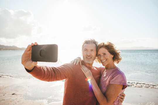 Happy mature woman with man taking selfie through smart phone at beach
