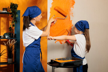 Mother and girl painting house walls with orange color paint, using renovating tools and paintbrush...