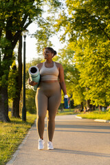 Plus sized African American woman walking with exercise mat and water through the park in a summer...