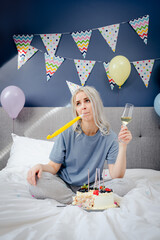 Sad, upset woman in pajama and party cap blowing in birthday pipe while sitting on bed with festive cake and champagne glass. Celebrates birthday alone. Bored party. Vertical card. Selective focus.