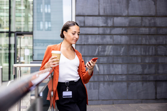 Businesswoman holding disposable cup looking at mobile phone by railing