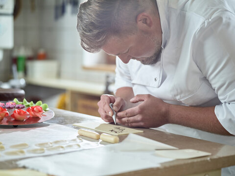 Confectioner Writing On Marzipan With Chocolate Icing At Kitchen