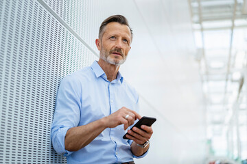 Thoughtful mature businessman with smart phone leaning on wall