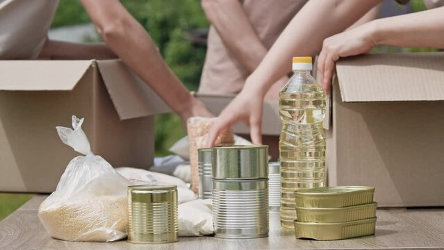 Close-up of unrecognizable members of charity organization pack canned and non-perishable products in cardboard boxes outdoors. Charity, donation and volunteering concept, global food crisis