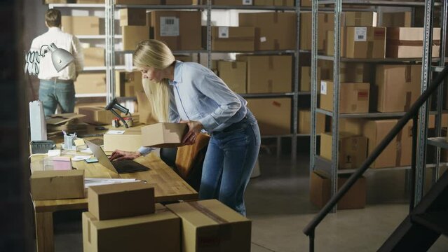 Inventory Manager Using Laptop Computer to Check Order Number on a Parcel, Preparing a Small Cardboard Box for Postage. Blond Female Small Business Owner Working in Warehouse with Colleague.