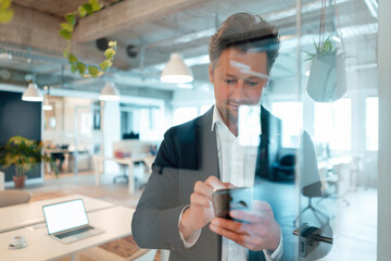 Mature businessman surfing net through mobile phone seen through glass in office