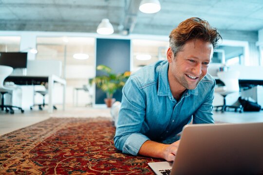 Happy Businessman Using Laptop Lying On Carpet In Office