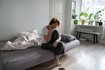 Depressed middle-aged woman sits on uncovered bed yawning after lonely sleepless night. Single...