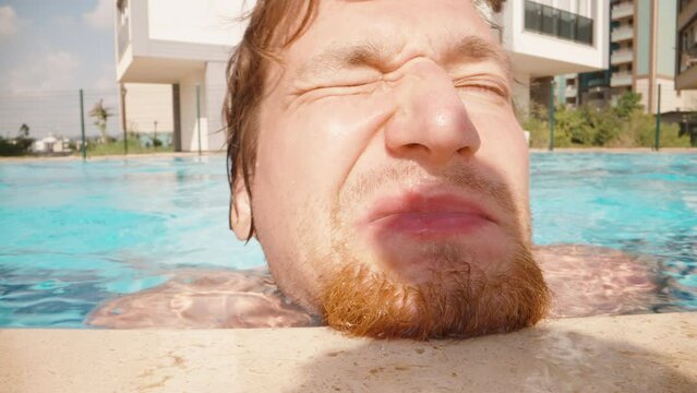 An unshaven man with a red beard in the pool, swims up to the camera and squints from the sun, close-up.