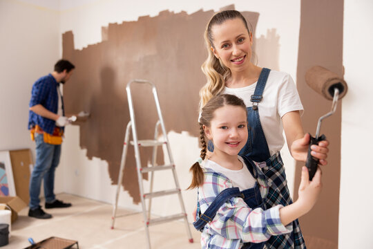 a beautiful blonde woman teaches her daughter how to paint the walls correctly. The girl is delighted with such an experience. Behind them is a father and a ladder.