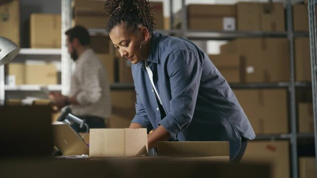 Inventory Manager Preparing a Small Cardboard Parcel for Postage. Multiethnic African American Female Small Business Owner Working on Laptop Computer in Warehouse. Handheld Footage.