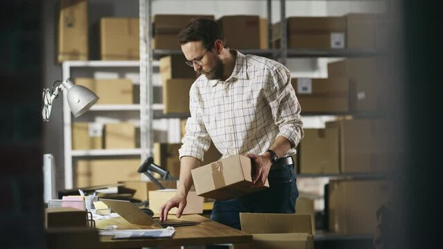 Small Business Owner of a Retail Online Shop Preparing a Small Cardboard Parcel for Postage. Stylish Young Inventory Manager Working on Laptop Computer in Warehouse Facility.