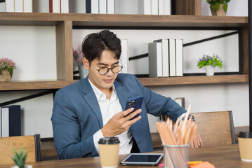 Portrait of young Asian business man in formal suit using smart phone in modern workplace.