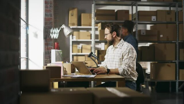 Male Inventory Manager Using Laptop Computer in Storeroom, Communicating with Clients, Writing Down Projects. Stylish Small Business Owner Working in Warehouse, Fulfilling Orders for Customers.