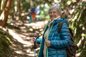 Happy active senior woman walking in the woods wearing backpack with a walking stick - caucasian...
