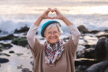 Happy caucasian senior woman at the beach at sunset light looking at camera making heart shape with...