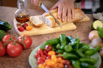 A rustic bread on a wooden cutting board and two woman's hands preparing a snack with a tomato bruschetta and spicy oil. Assortment of fresh vegetables on the table