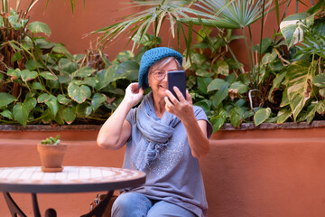 Smiling mature woman sitting at coffee shop looking at herself in mobile phone adjusting her cap