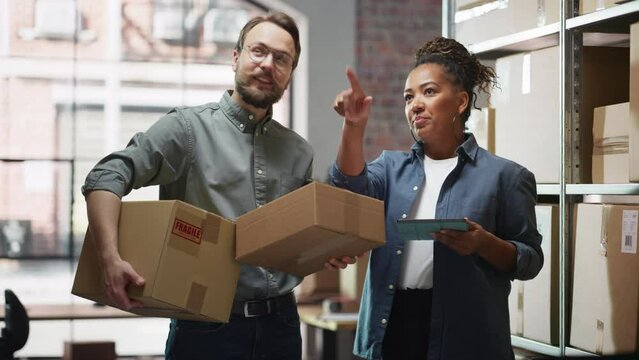 Female Inventory Manager Using Tablet Computer, Talking to a Worker Holding Two Cardboard Packages. They Discuss Customer Orders. Stock of Parcels with Products Ready for Shipment in the Background.