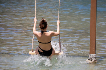 Woman in black swimsuit ride on a swing set in sea water. Tourist having fun on a beach, vacation in summer