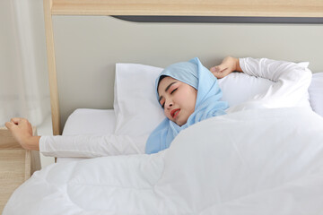 Obraz na płótnie Canvas Beautiful asian woman wearing white muslim sleepwear lying on bed, stretching her arms after getting up in the morning at sunrise. Cute young girl with hijab wake up and relaxing, closing her eyes