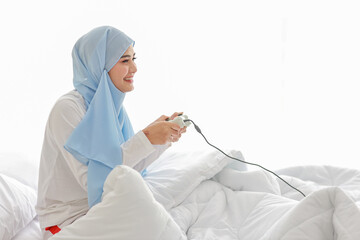 VDO game console station concept. active asian woman wearing white muslim sleepwear sitting on bed, holding joystick and playing exciting game. Cute girl looked excited with game controller console