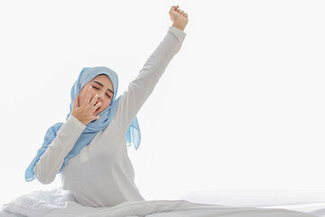 Beautiful asian woman stretching her arms after getting up in the morning on bed at sunrise in bedroom. Cute young girl in muslim dress and hijab sitting and relaxing with happy and smiling face.