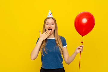 Young cheerful amazed woman in party cone holds red inflated helium balloon, celebrates birthday or...