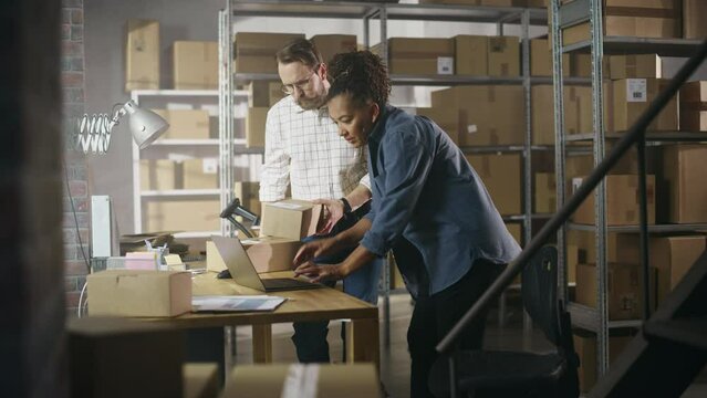 Diverse Male and Female Warehouse Inventory Managers Talking, Using Laptop Computer and Checking Retail Stock. Rows of Shelves Full of Cardboard Box Packages in the Background.