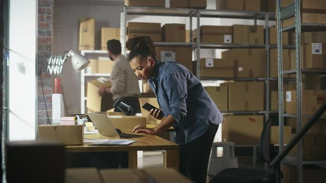 Inventory Manager Using Smartphone to Scan a Barcode on Parcel, Preparing a Small Cardboard Box for Postage. Black Multiethnic Small Business Owner Working on Laptop in Warehouse with Colleague.