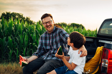 father and son sits on truck of car and eating watermelon. they are outdoor in corn field