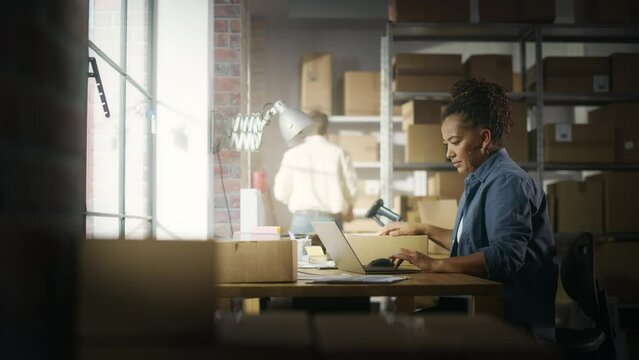 Black Female Warehouse Inventory Manager Works on Laptop Computer while Sitting at Her Desk. In the Background Employee Working in the Room Full of Cardboard Box Parcels Ready For Shipping.