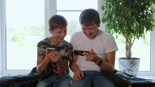 Two boys play computer games at home on their smartphones, watch videos, rejoice and laugh. Two brothers watch interesting videos together on tablet or smartphone. They laugh and indulge. Sit on table