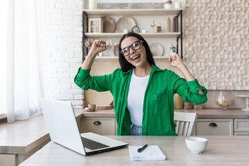 Portrait of happy young business woman in glasses and green jacket. She sits at the table at home, works with a laptop, is happy, raised her hands up, received good news, smiles.