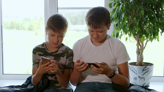 Two boys play computer games at home on their smartphones, watch videos, rejoice and laugh. Two brothers watch interesting videos together on tablet or smartphone. They laugh and indulge. Sit on table