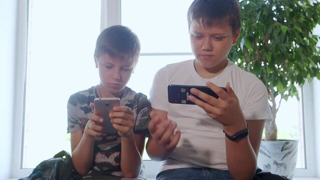 Two boys play computer games at home on their smartphones, watch videos, rejoice and laugh. Two brothers watch interesting videos together on tablet or smartphone. They laugh and indulge. Sit on table