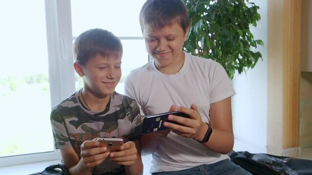 Two boys play computer games at home on their smartphones, watch videos, rejoice and laugh. Two brothers watch interesting videos together on tablet or smartphone. They laugh and indulge. Sit on table