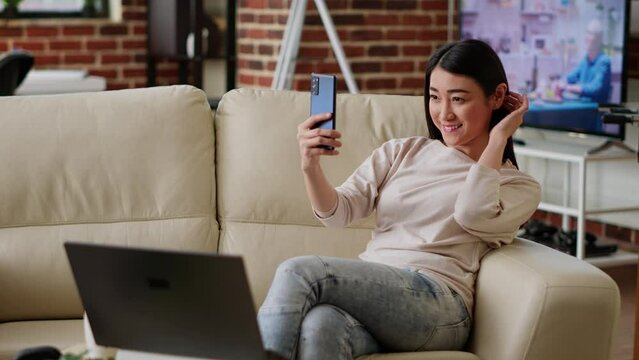 Smiling heartily woman taking selfie photos with smartphone while working remotely from home. Joyful person photographing herself with modern touchscreen phone device while doing remote work