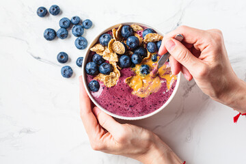 Blueberry smoothie bowl with peanut butter, cereal and fresh berries, white marble background.