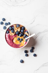 Blueberry smoothie bowl with peanut butter, cereal and fresh berries, white marble background.