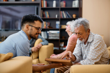 Elderly man uses touchpad with help of young healthcare worker at nursing home.