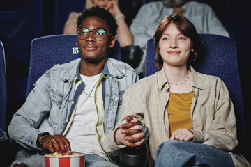 Joyful African American man and Caucasian woman in love having date at cinema watching movie and holding hands