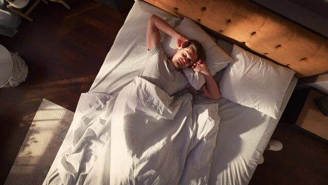 Top View Home: Handsome Young Man Sleeps in Her Bed, Sun Shines on Him, He Opens Eyes and Greets New Sunny Day at Home. Joyful Waking Up of a Guy who is Ready for Productive Day. Top Down Above Shot