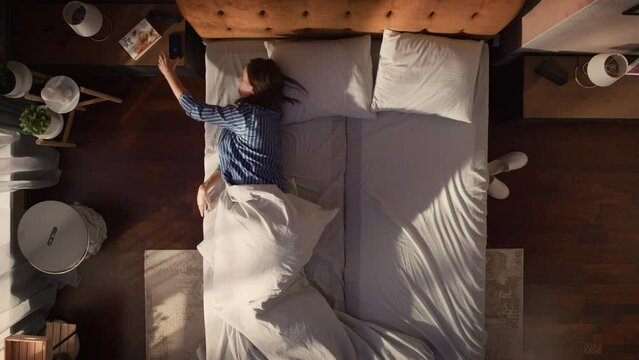 Top View Apartment: Beautiful Young Woman Sleeps Charmingly in Her Bed, Turns off Smartphone Alarm Clock, Greets a New Day with Looking up at Camera and Smiling. Top Down