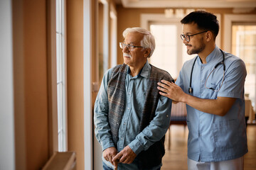Male caregiver and senior man look through the window ar nursing home.