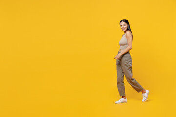 Full body smiling happy fun cool young latin woman 30s she wearing basic beige tank shirt walk stroll going look camera isolated on plain yellow backround studio portrait. People lifestyle concept