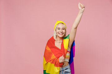 Young confident fun blond lesbian woman she wear yellow hat wrapped in striped flag do hero gesture clench fist isolated on plain pastel light pink background. People lgbtq lifestyle fashion concept.