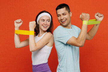 Young smiling trainer instructor sporty two man woman in headband t-shirt hold using hands fitness elastic bands spend weekend in home gym isolated on plain orange background. Workout sport concept.