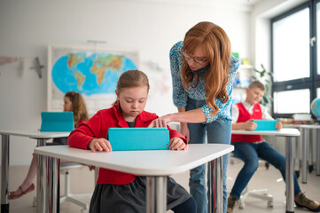 Down syndrome schoolgirl using tablet with help of teacher during class at school, integration...
