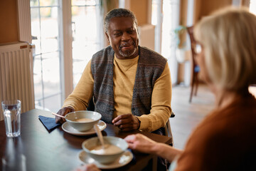 Happy black senior communicating with female friend while eating lunch in nursing home.